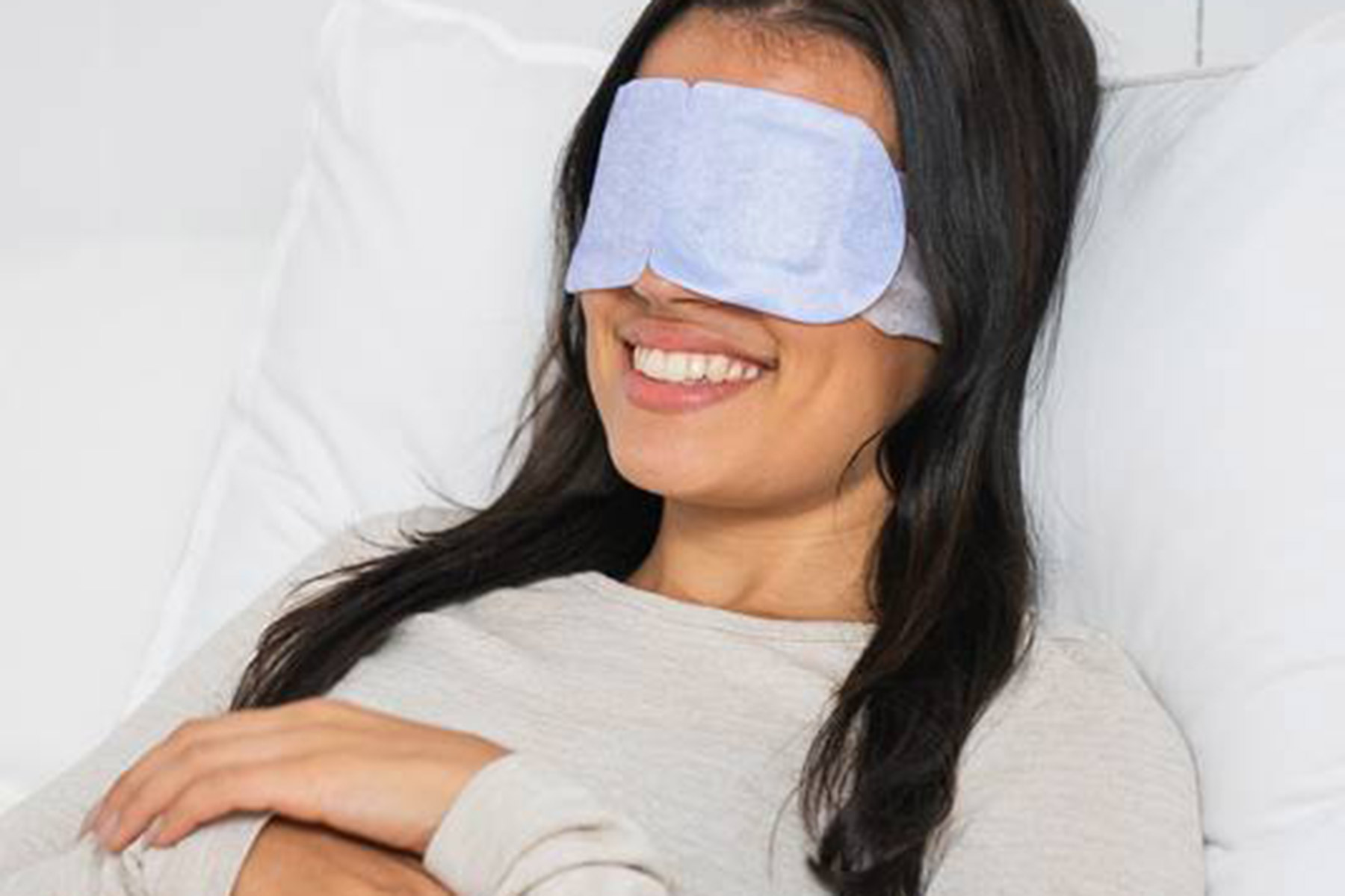 A woman with long dark hair is lying on a bed, smiling, and wearing a light blue eye mask. She is dressed in a light-colored long-sleeve shirt and appears relaxed and comfortable. Fearrington Village