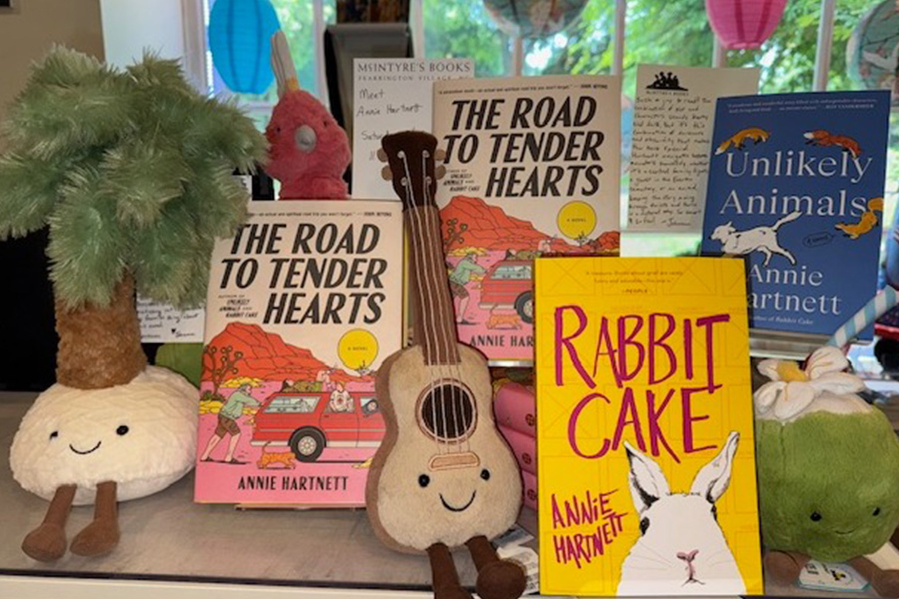 Gentle Reader - May 20, 2025 1 A display of three books by Annie Hartnett—“The Road to Tender Hearts,” “Rabbit Cake,” and “Unlikely Animals”—arranged among plush toys and a small toy guitar, with a leafy tree visible outside the window. Fearrington Village