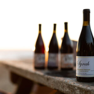 Five wine bottles are arranged on a rustic wooden table outdoors at sunset, with two bottles labeled "Syrah" in the foreground and the ocean out of focus in the background. Fearrington Village