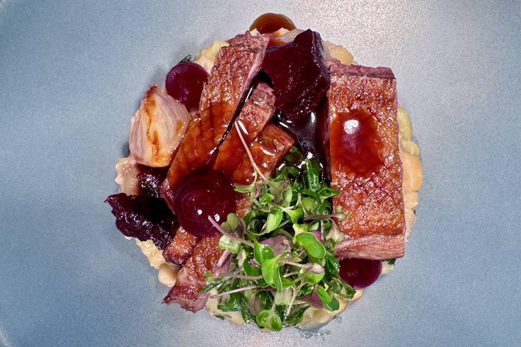 A plated gourmet dish featuring seared duck breast, microgreens, beans, glazed onions, red beets, and a glossy brown sauce, all artfully arranged on a gray plate. Fearrington Village