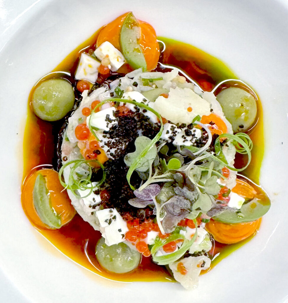A vibrant gourmet dish topped with microgreens and herbs, surrounded by sliced tomatoes, green grapes, orange oil, fish roe, and cubes of soft cheese on a white plate. Fearrington Village