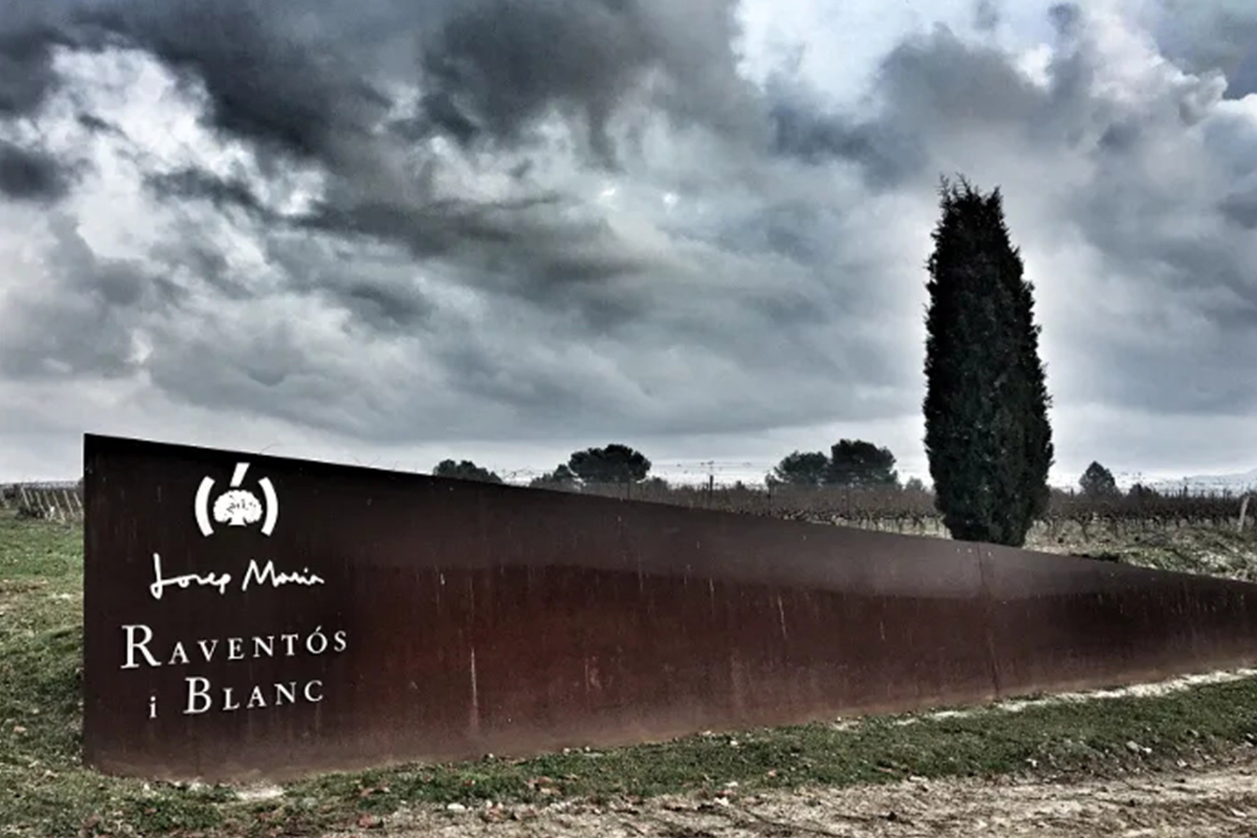 A rust-colored metal sign reading "Josep Muir RAVENTÓS i BLANC" stands beside a tall cypress tree under a dramatic, cloudy sky in a vineyard landscape. Fearrington Village