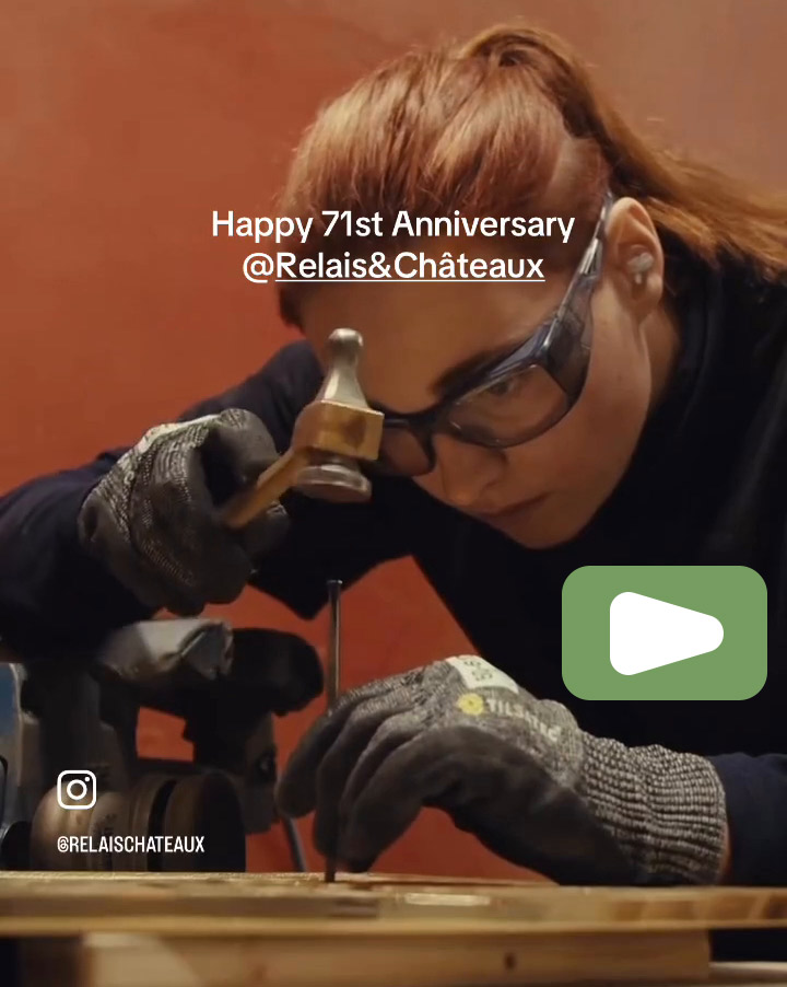 A woman with red hair and safety glasses uses a hammer and nail on a wooden surface. Text reads "Happy 71st Anniversary @Relais&Châteaux." She is focused and wearing gloves in a workshop setting. Fearrington Village