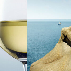 A glass of white wine on the left is visually aligned with a coastal rock formation and the ocean on the right, creating an artistic blend of the wine and sea. A small sailboat is visible on the horizon. Fearrington Village