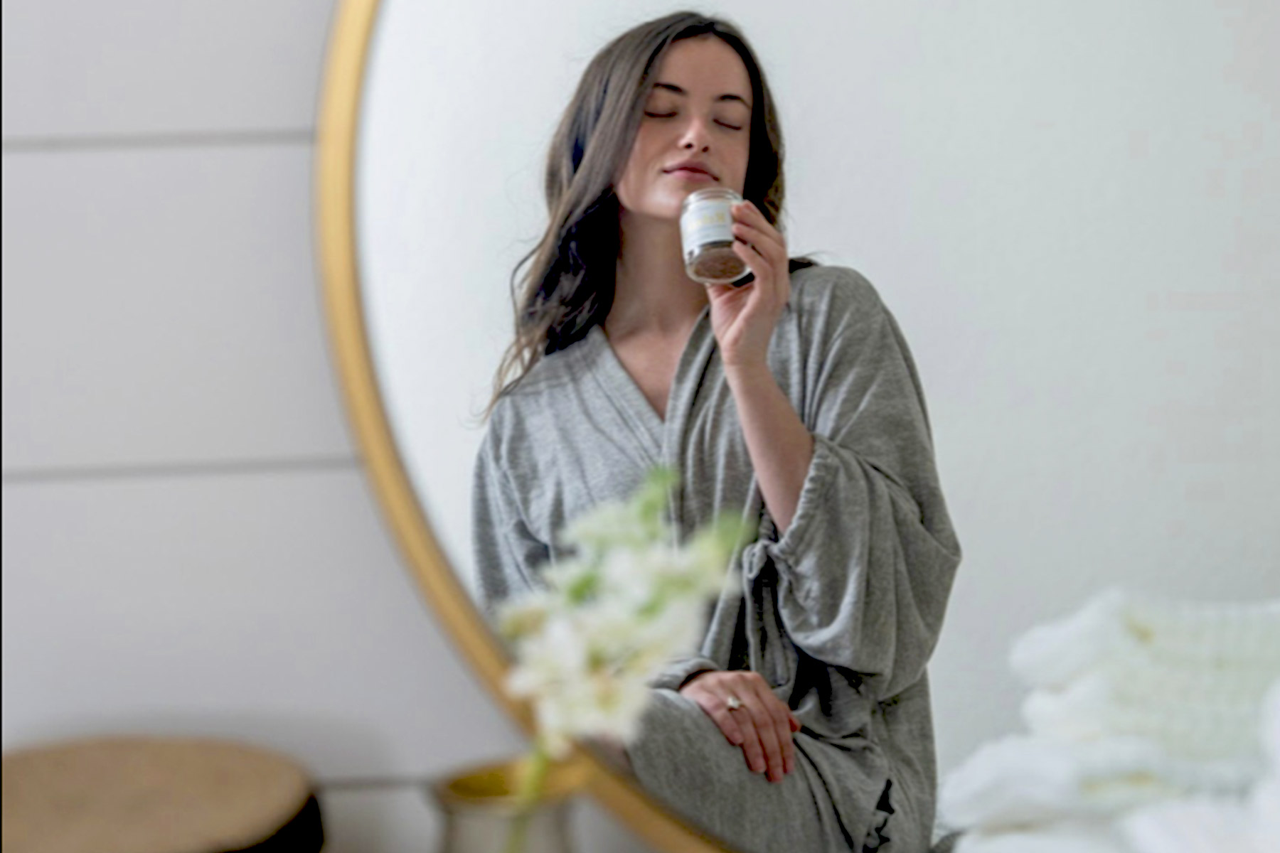 The Spa Edit - We're Expanding! 1 A woman in a grey robe sits in front of a round mirror, holding a small jar near her face with a relaxed expression. A blurred plant and stacked towels are visible in the foreground. Fearrington Village