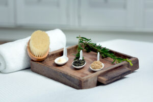 A wooden tray holds three white ceramic spoons with different powders and herbs, a sprig of rosemary, a rolled white towel, and a dry body brush on a white surface, suggesting a spa or wellness setting. Fearrington Village