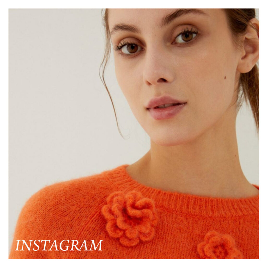 A woman in an orange sweater with knitted flower details looks at the camera. The image has a soft background and the word "INSTAGRAM" appears in white text at the bottom left corner. Fearrington Village