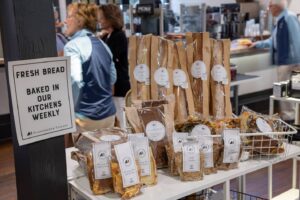 A display of packaged bread and snacks sits on a white table in a bakery. A sign reads, “Fresh bread baked in our kitchens weekly.” People are seen talking and working in the background. Fearrington Village