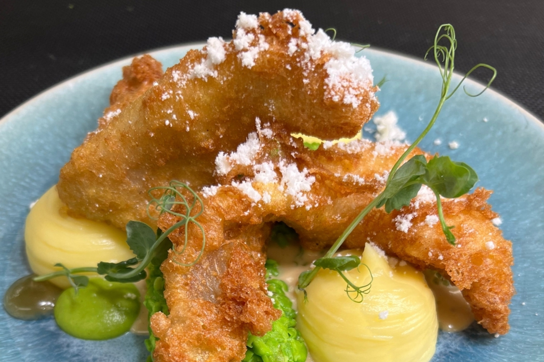 Golden battered fish fillets are artfully arranged on a blue plate with creamy mashed potatoes, green pea purée, microgreens, and a dusting of white powder for garnish. Fearrington Village