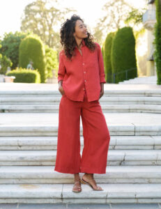 A woman stands on marble steps outdoors, wearing a loose-fitting, long-sleeve red shirt and matching wide-leg pants. She has curly hair, hands in her pockets, and gazes to the side, smiling. Greenery and trees are in the background. Fearrington Village