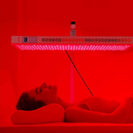 A person lying on their back under a rectangular panel emitting red light, likely undergoing red light therapy, in a room illuminated by the red glow. Fearrington Village