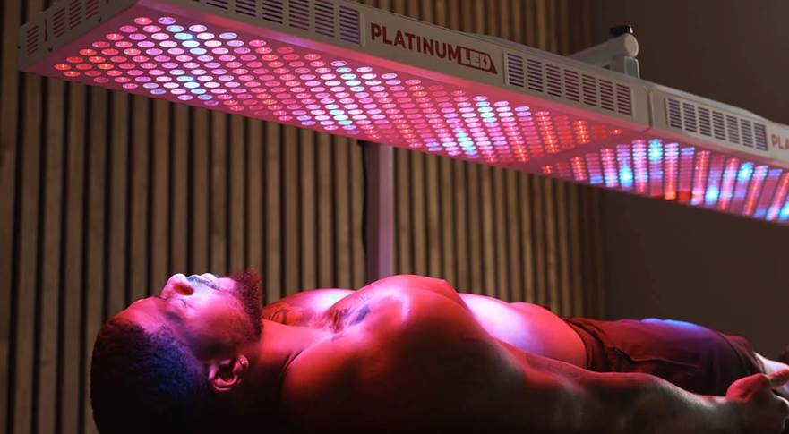 A muscular man lies on his back under a large red and near-infrared LED light therapy panel marked "PLATINUM LED" in a modern room with wooden walls. Fearrington Village