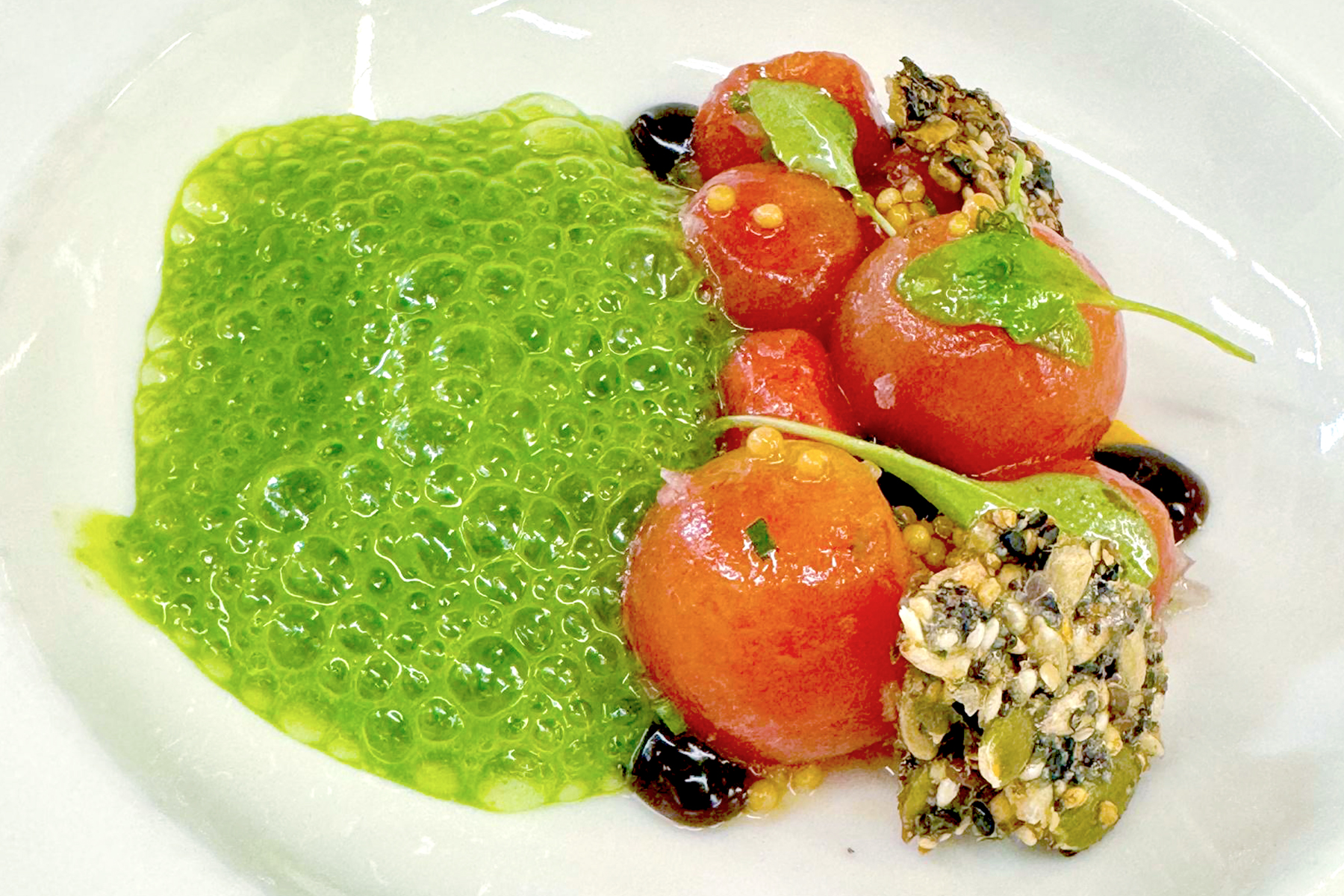 A gourmet dish featuring bright green bubbly sauce, several peeled cherry tomatoes, and a crunchy seed-coated garnish, all artfully arranged on a white plate. Fearrington Village