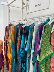 A clothing rack displays colorful, patterned garments in shades of blue, green, yellow, and red, hanging neatly on white hangers under a sign that reads "GIGEEZ. Fearrington Village