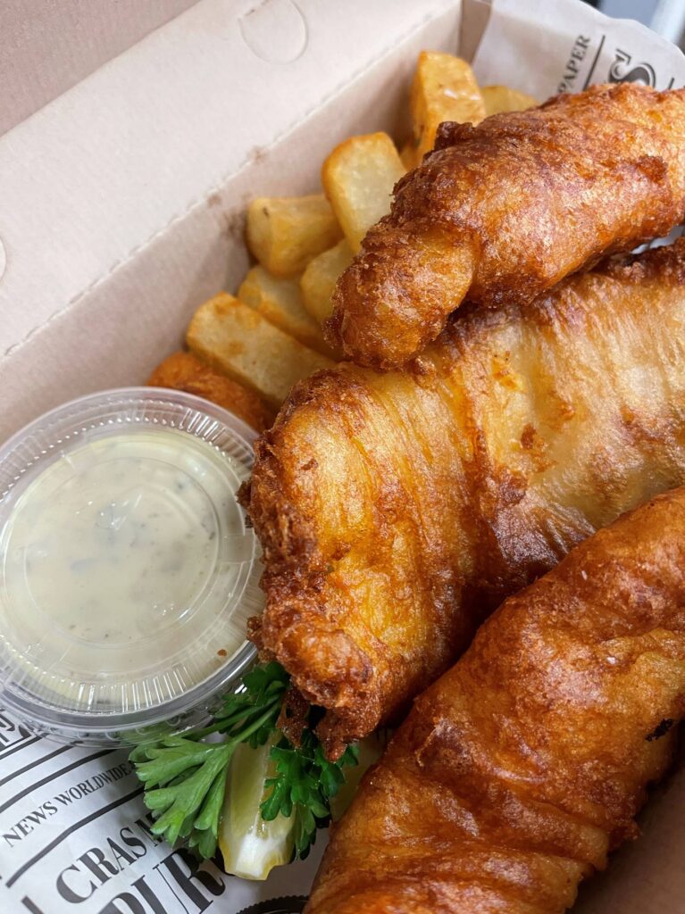 Golden fried fish fillets with thick-cut fries in a cardboard box, served with a small cup of tartar sauce, a lemon wedge, and a sprig of parsley, on a newspaper-style liner. Fearrington Village