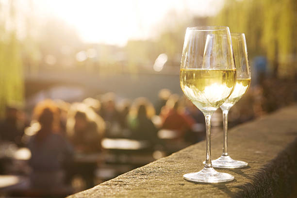 Two glasses of white wine sit on a stone ledge in the foreground, with a group of people sitting and socializing in an outdoor setting on a sunny day, blurred in the background. Fearrington Village