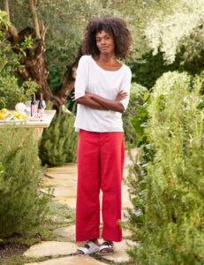 A woman with curly hair stands outdoors on a stone path, surrounded by greenery. She wears a white top, red wide-leg pants, and white sandals. A table with drinks and fruit is visible in the background. Fearrington Village