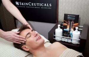 A man is lying on a spa bed with his eyes closed, receiving a facial massage. Bottles of SkinCeuticals skincare products are displayed nearby, and a SkinCeuticals sign is in the background. Fearrington Village
