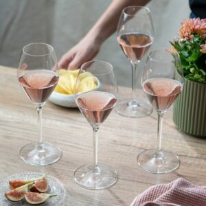 Four glasses of rosé wine on a wooden table with a plate of sliced figs, a bowl of potato chips, a pink cloth, and a green potted plant. A person's hand reaches for the chips in the background. Fearrington Village