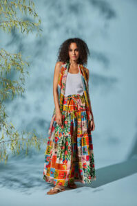 A woman wearing a white tank top and a colorful, patchwork-patterned maxi skirt with a matching scarf stands on a light blue background next to leafy branches. She has curly hair and is wearing sandals. Fearrington Village