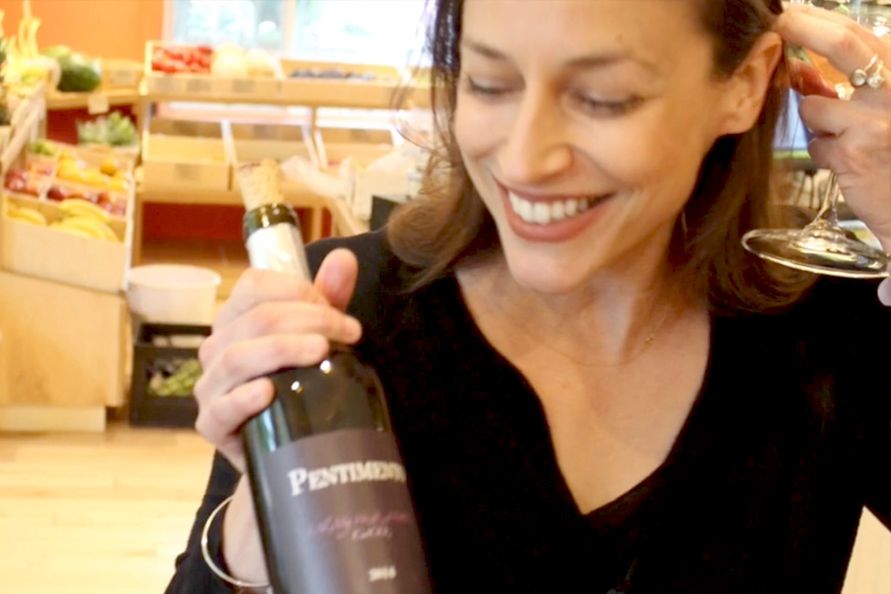 A smiling woman in a black top holds a bottle of Pentimento red wine and a wine glass in a warmly lit room with shelves of produce in the background. Fearrington Village