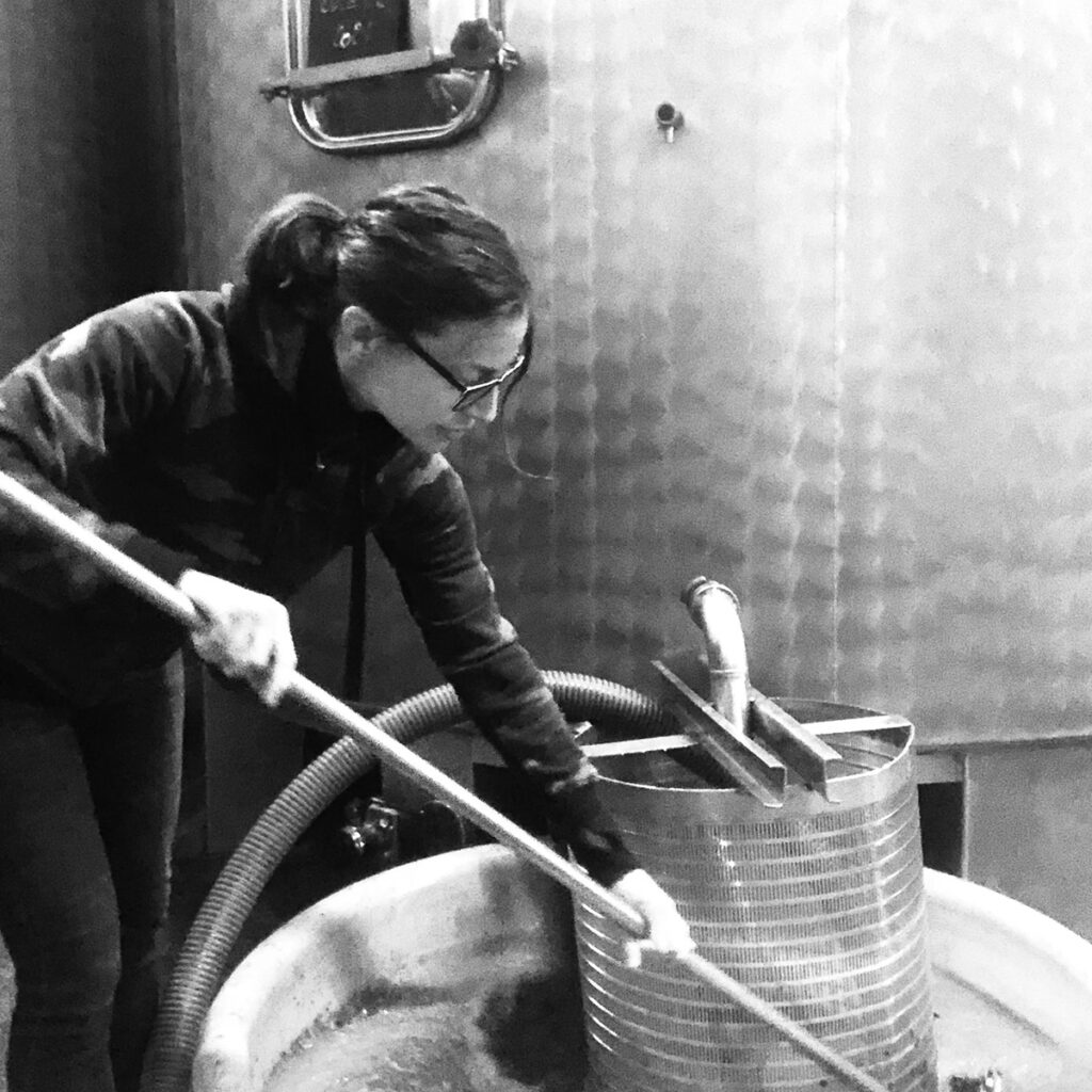 A person wearing glasses stirs liquid in a large vat with a long pole inside an industrial setting, possibly a brewery or winery, next to a tall metal tank. The image is in black and white. Fearrington Village