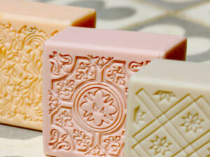 Three decorative soap bars in peach, pink, and cream colors, each with intricate embossed floral and geometric patterns, placed on a tiled surface. Fearrington Village