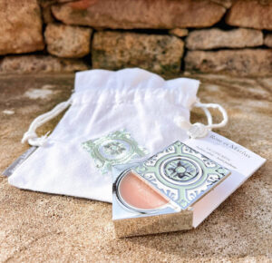 A decorative solid perfume tin with intricate patterns rests on a sandy surface, partially opened, beside a white drawstring pouch embroidered with a matching design. Rustic stone blocks are in the background. Fearrington Village