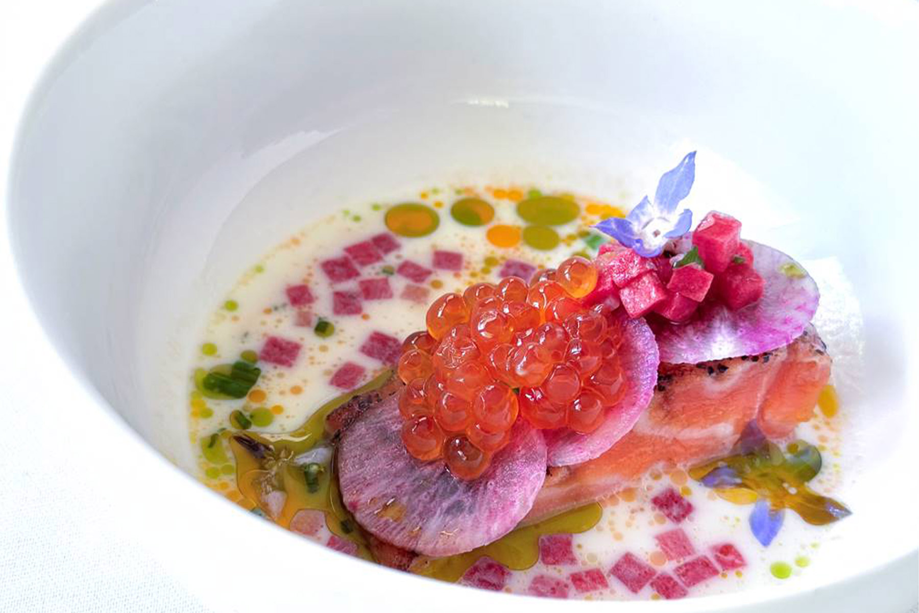 A gourmet dish featuring a slice of salmon topped with salmon roe, thin radish slices, diced vegetables, edible flowers, and colorful oil drops, beautifully arranged in a white bowl with a light sauce. Fearrington Village
