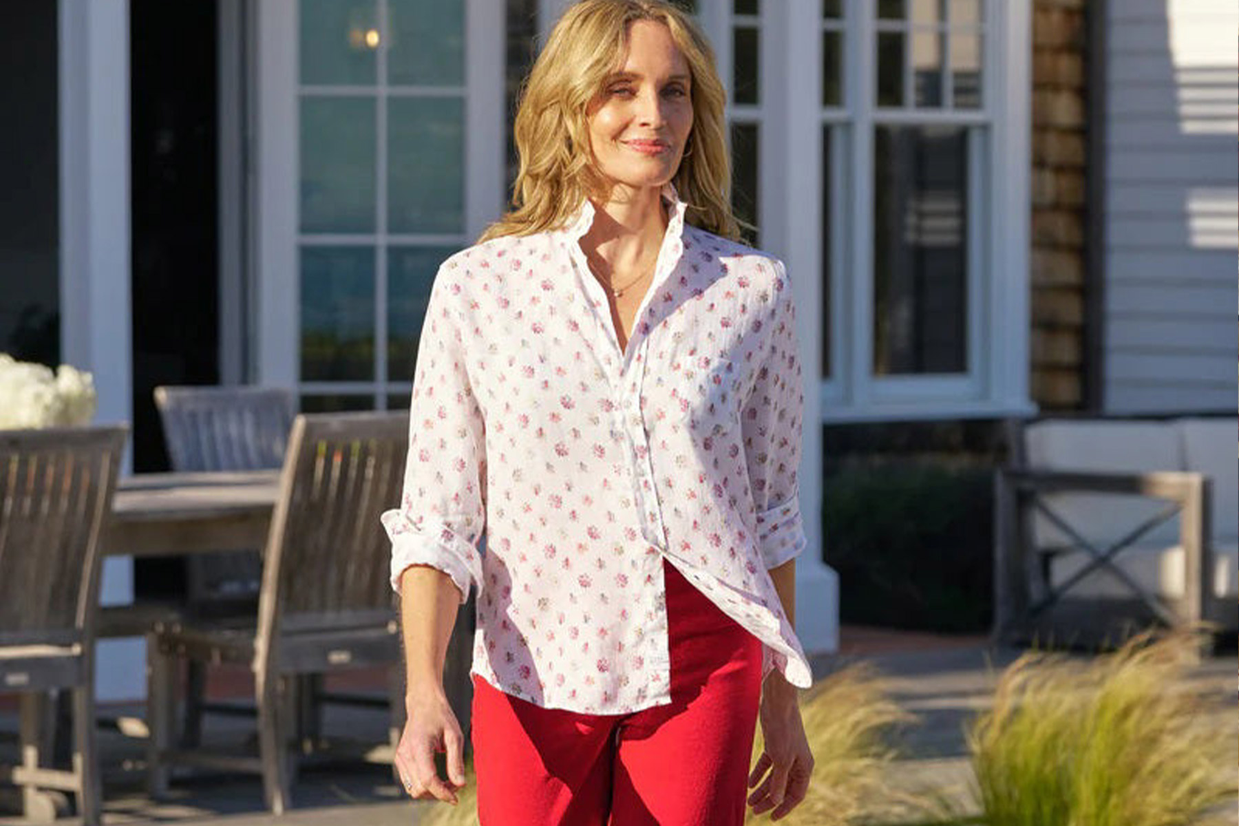 A woman with blonde hair wearing a white button-up shirt with a small pattern and red pants walks outside near a patio with wooden furniture and large windows in the background. Fearrington Village