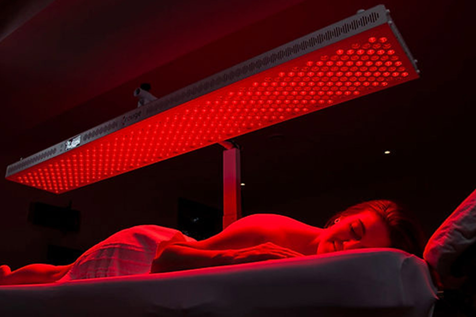 A person lies face down on a massage table, covered with a towel, under a large panel emitting red light in a dark room, suggesting a red light therapy session. Fearrington Village
