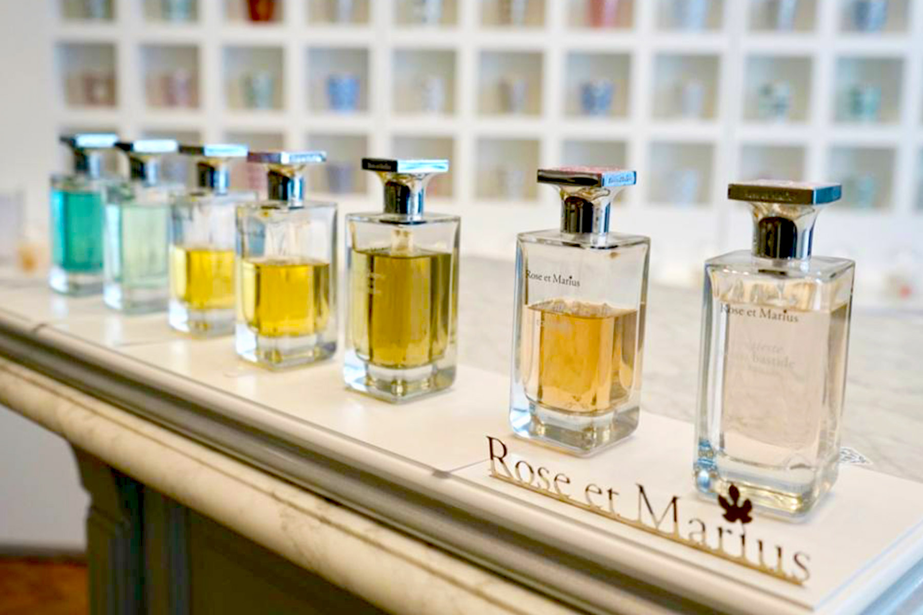A row of assorted Rose et Marius perfume bottles with varying colors of liquid sits on a marble countertop, with a white display and shelves in the blurred background. Fearrington Village