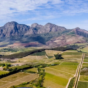 Aerial view of a patchwork of green and yellow fields with dirt roads, set against a backdrop of rugged mountains under a partly cloudy blue sky. Fearrington Village