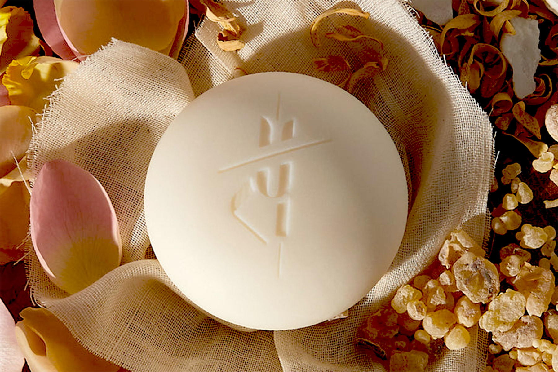 A round, off-white soap bar with a minimalistic design is placed on sheer fabric, surrounded by dried flower petals and pieces of frankincense resin. Fearrington Village