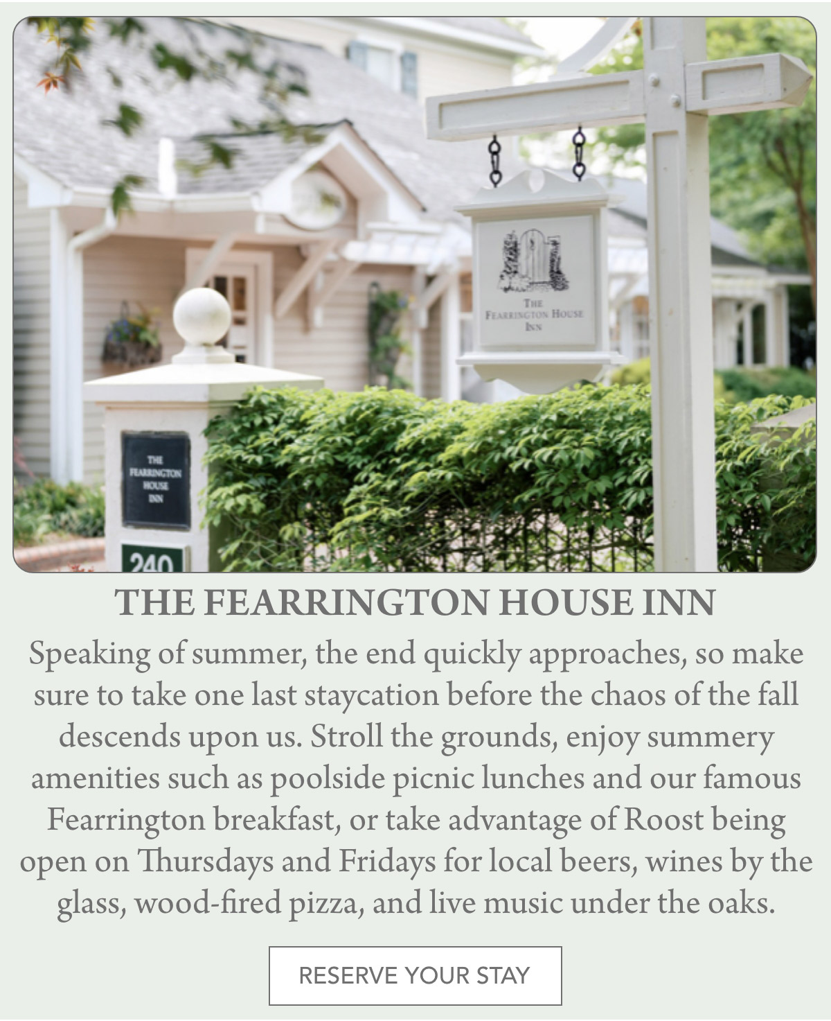 A white sign for The Fearrington House Inn hangs on a post outside a charming building with gray siding and white trim, surrounded by lush greenery in a tranquil setting. Fearrington Village