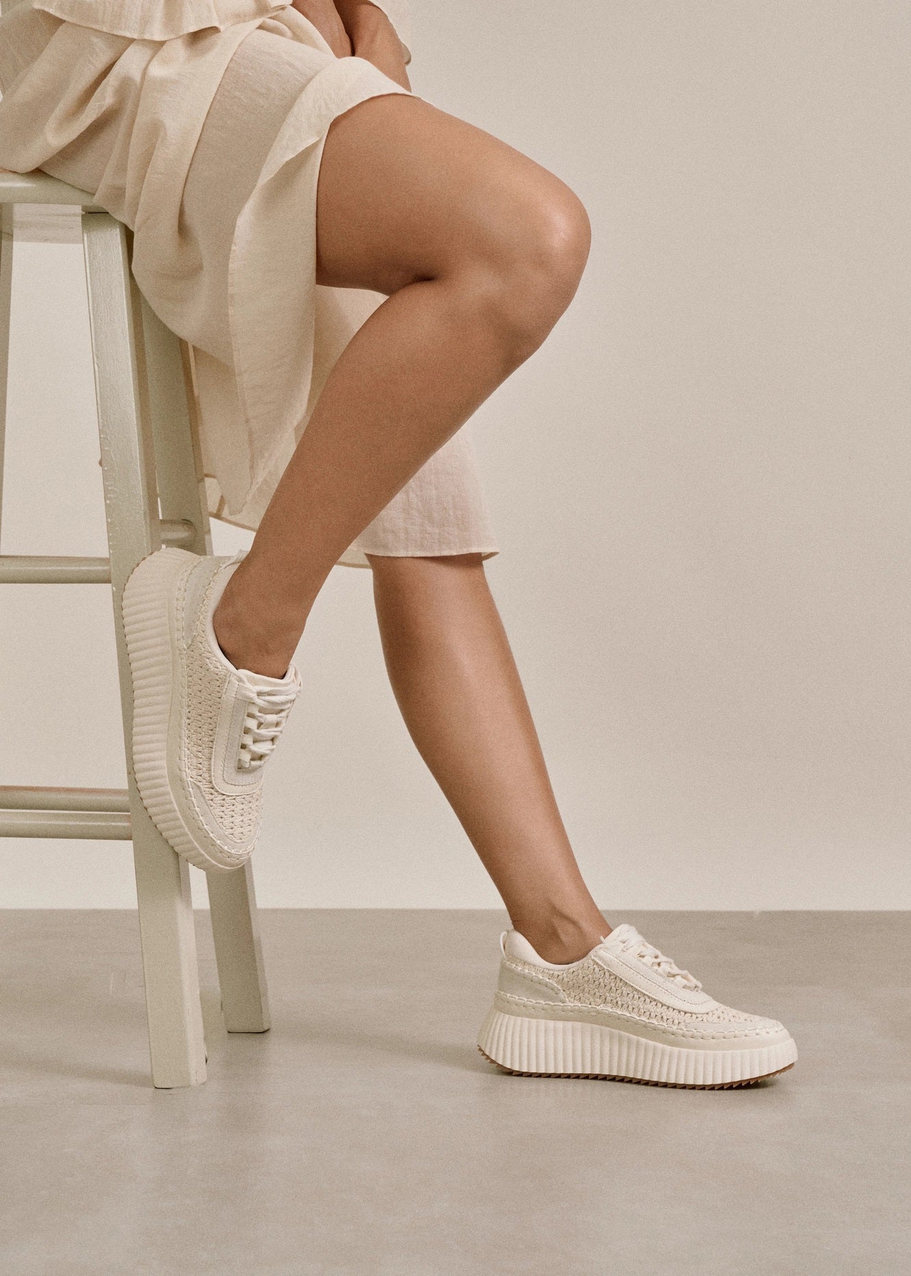 A person sits on a light-colored stool, wearing a cream-colored skirt and white platform sneakers with textured details, against a neutral background. Fearrington Village
