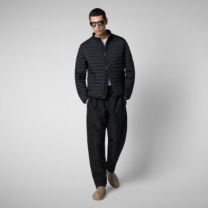 A man wearing sunglasses, a black quilted jacket, wide-leg black pants, and tan slip-on shoes stands against a plain light gray background with his hands in his pockets. Fearrington Village