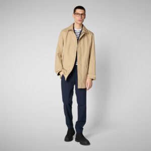 A man wearing glasses, a beige trench coat, a navy and white striped shirt, navy pants, and black shoes stands against a plain light gray background. Fearrington Village