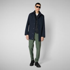 A man wearing sunglasses, a dark navy coat over a white shirt, green pants, and black shoes stands against a plain light gray background. Fearrington Village