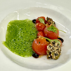 A gourmet dish featuring peeled cherry tomatoes, a green foamy sauce, black and white seed clusters, microgreens, and dark balsamic dots, artistically arranged on a white plate. Fearrington Village