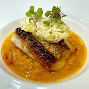 A seared fish fillet with crispy skin sits on orange sauce, topped with a dollop of mashed potatoes and microgreens, presented on a white plate. Fearrington Village