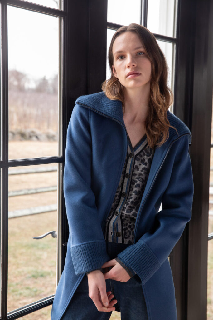 A woman with wavy brown hair stands indoors by large windows, wearing a blue coat over a leopard print cardigan and blue jeans. She looks at the camera with a neutral expression. Fearrington Village
