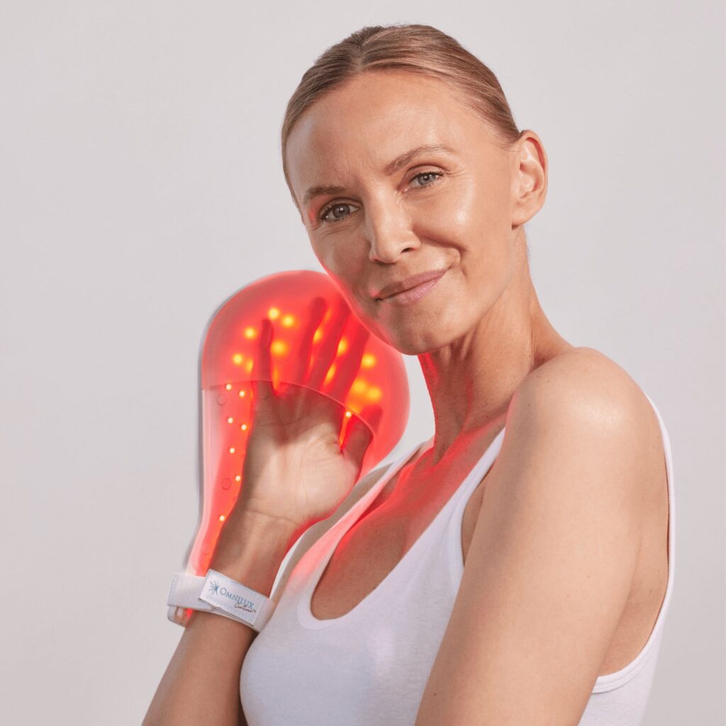 A woman in a white tank top smiles while holding a glowing red LED light therapy device to her face. The device emits multiple small lights and is secured to her hand with a strap. Fearrington Village