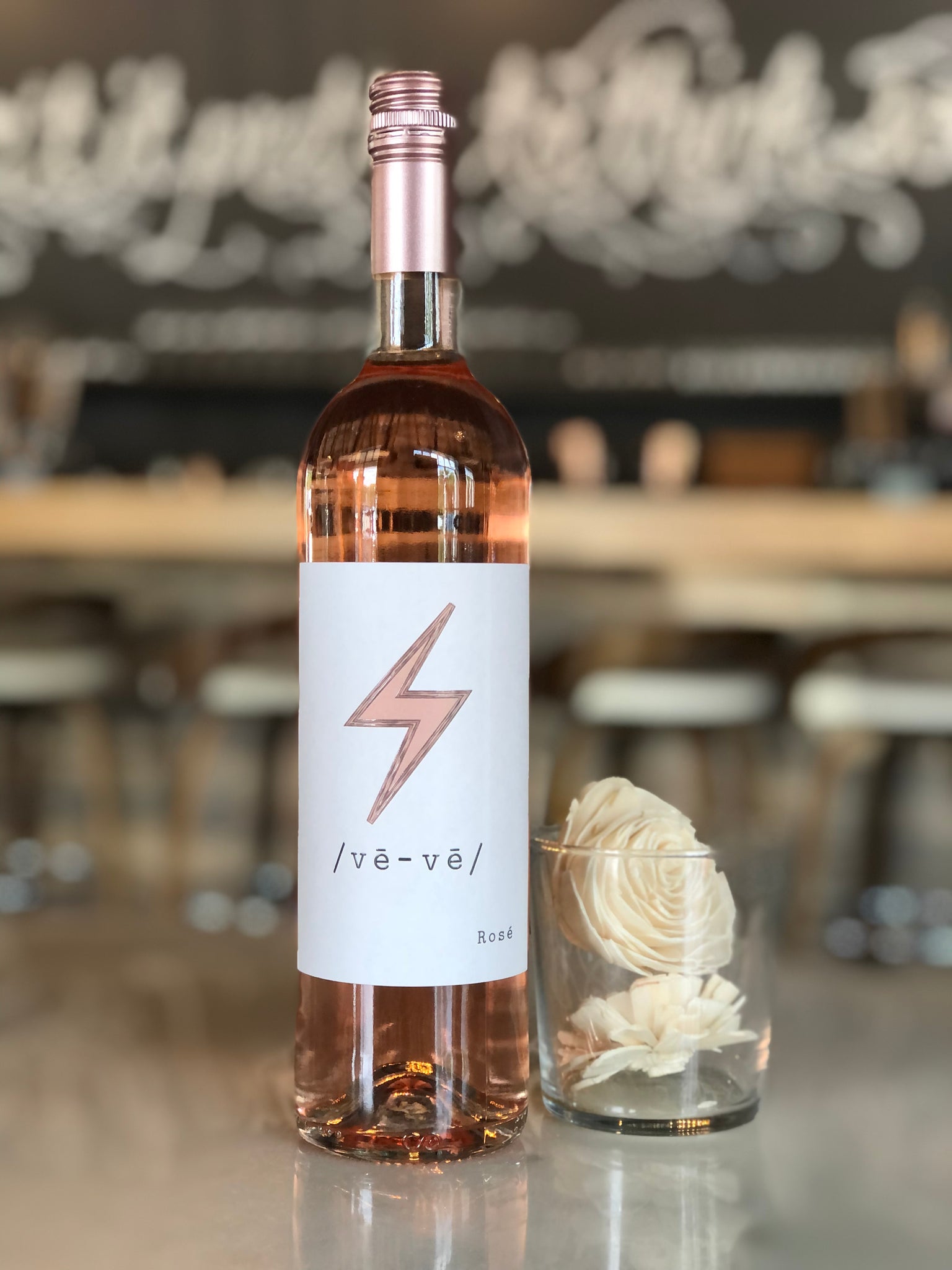 A bottle of rosé wine with a lightning bolt label sits on a reflective table beside a glass filled with cream-colored flowers; a blurred bar background is visible. Fearrington Village