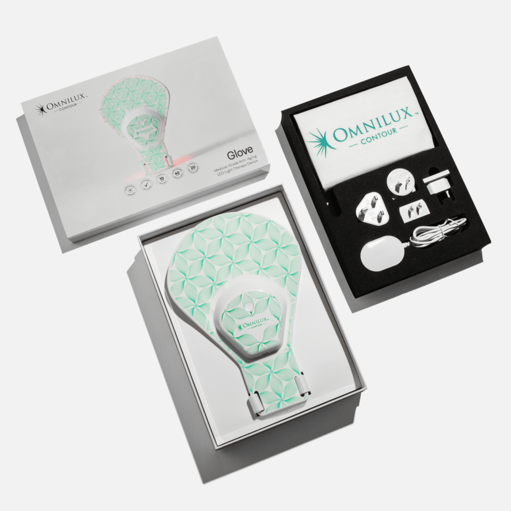 Flat lay of the Omnilux Contour Glove set, showing the glove-shaped LED device in its box, an open accessory tray with charging cables, plugs, controller, and a branded cloth, and the product packaging. Fearrington Village