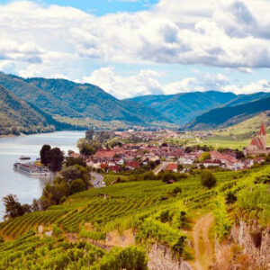 A picturesque riverside village with red-roofed buildings sits beside a winding river, surrounded by lush green vineyards and hills under a partly cloudy sky. A riverboat is docked by the village. Fearrington Village