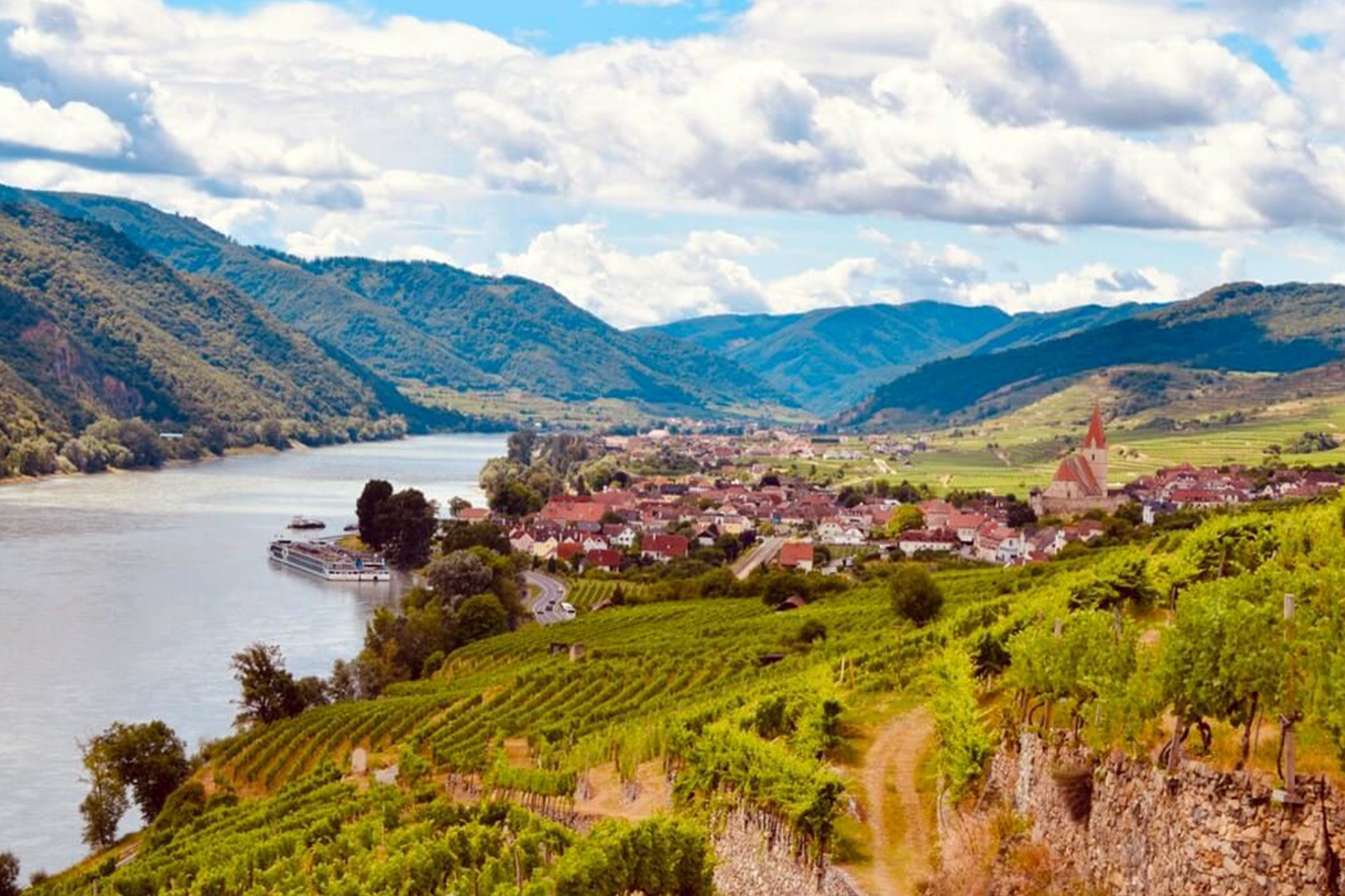 A picturesque riverside village with red-roofed buildings sits beside a winding river, surrounded by lush green vineyards and hills under a partly cloudy sky. A riverboat is docked by the village. Fearrington Village