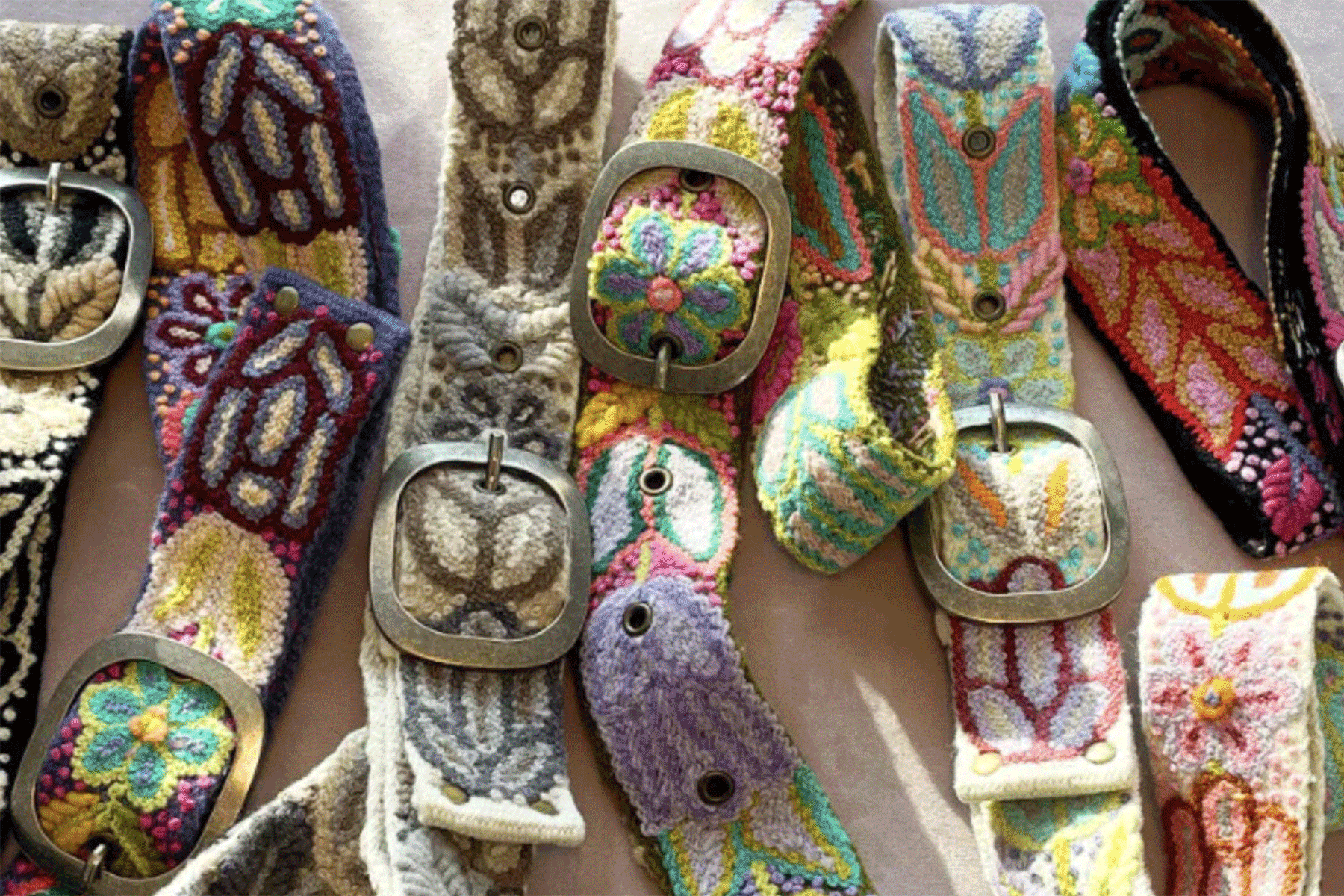 A collection of colorful, embroidered belts with ornate metal buckles are displayed side by side, featuring floral and abstract patterns in various vibrant shades. Fearrington Village