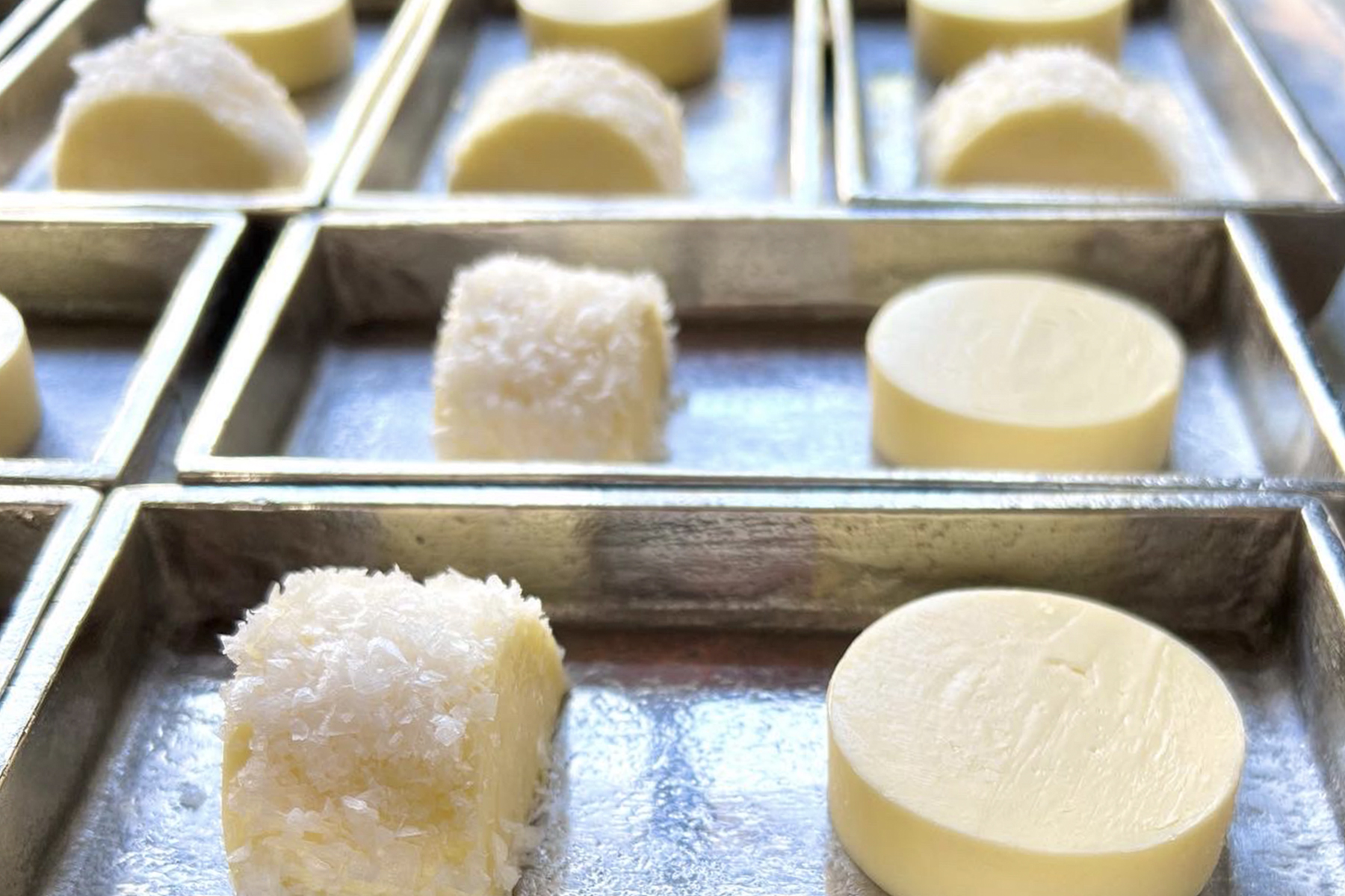 Close-up of metal trays holding round yellow sweets, some plain and some coated with shredded coconut, arranged in neat rows with sunlight highlighting their smooth and textured surfaces. Fearrington Village