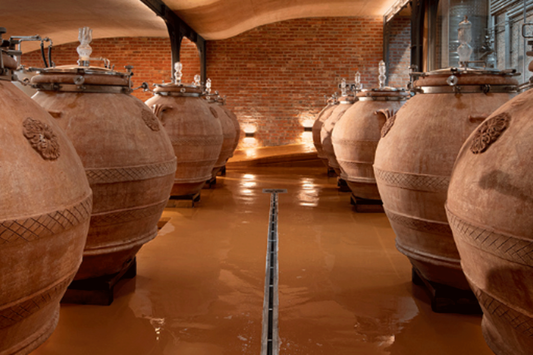 Rows of large, round clay amphorae with decorative patterns are lined up on a shiny floor in a brick-walled, warmly lit cellar, used for fermenting or storing wine or other beverages. Fearrington Village